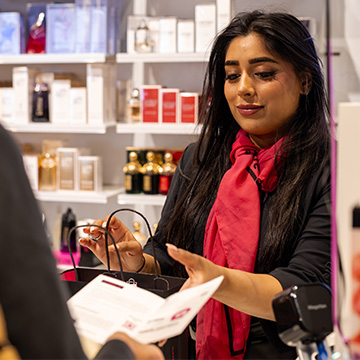 Collaborateur d’ICI PARIS XL remettant un achat à un client à la caisse, avec des parfums et produits de beauté en arrière-plan.