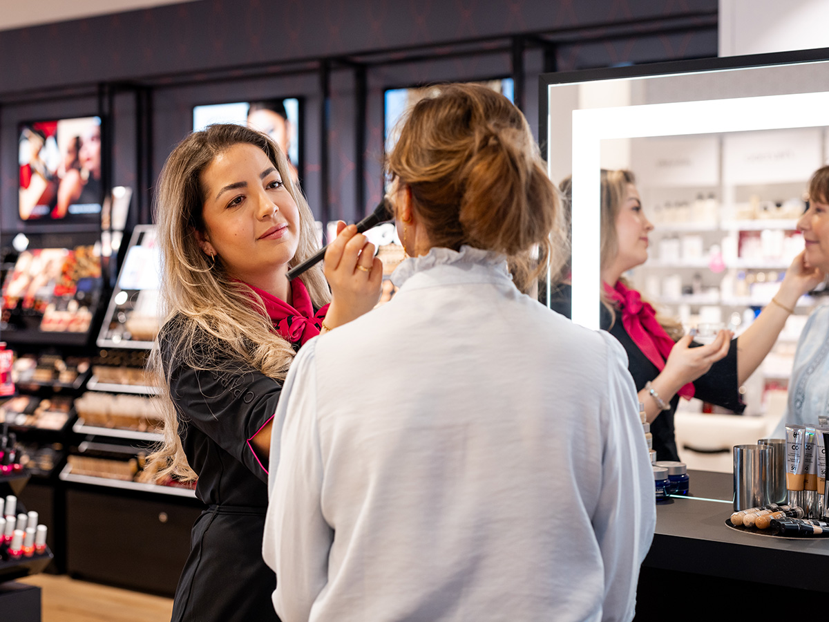 Conseiller en magasin ICI PARISXL appliquant du maquillage à un client devant un miroir éclairé, entouré de produits cosmétiques.