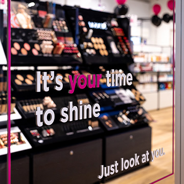 Miroir dans un magasin ICI PARIS XL affichant le message « It's your time to shine », avec des rayons de maquillage en arrière-plan.