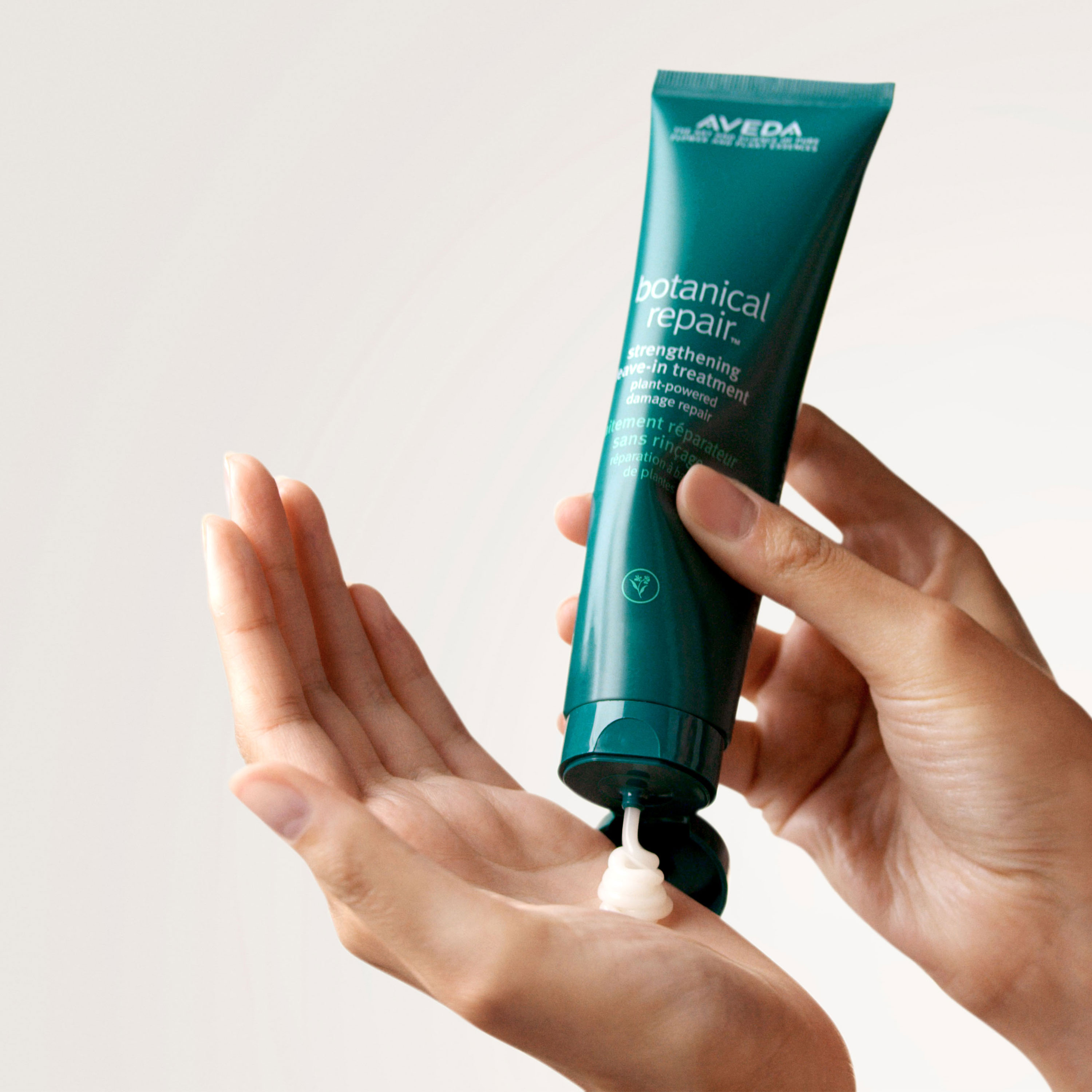 A hand holding the Aveda botanical repair leave-in treatment and other hand squeezing the product.