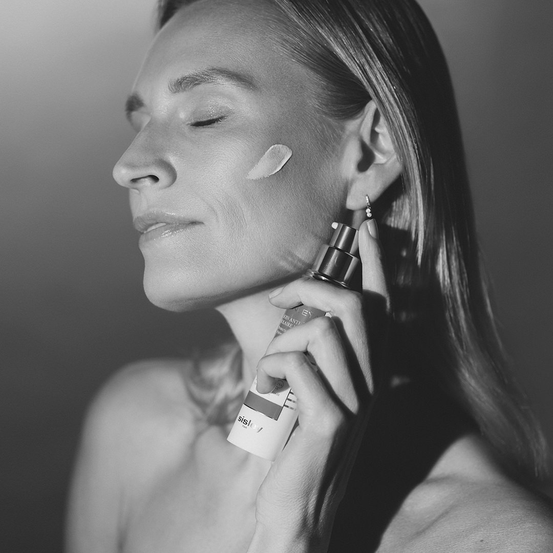 Black and white photo of a woman applying Sisley Sunleÿa cream with a visible dab on her cheek