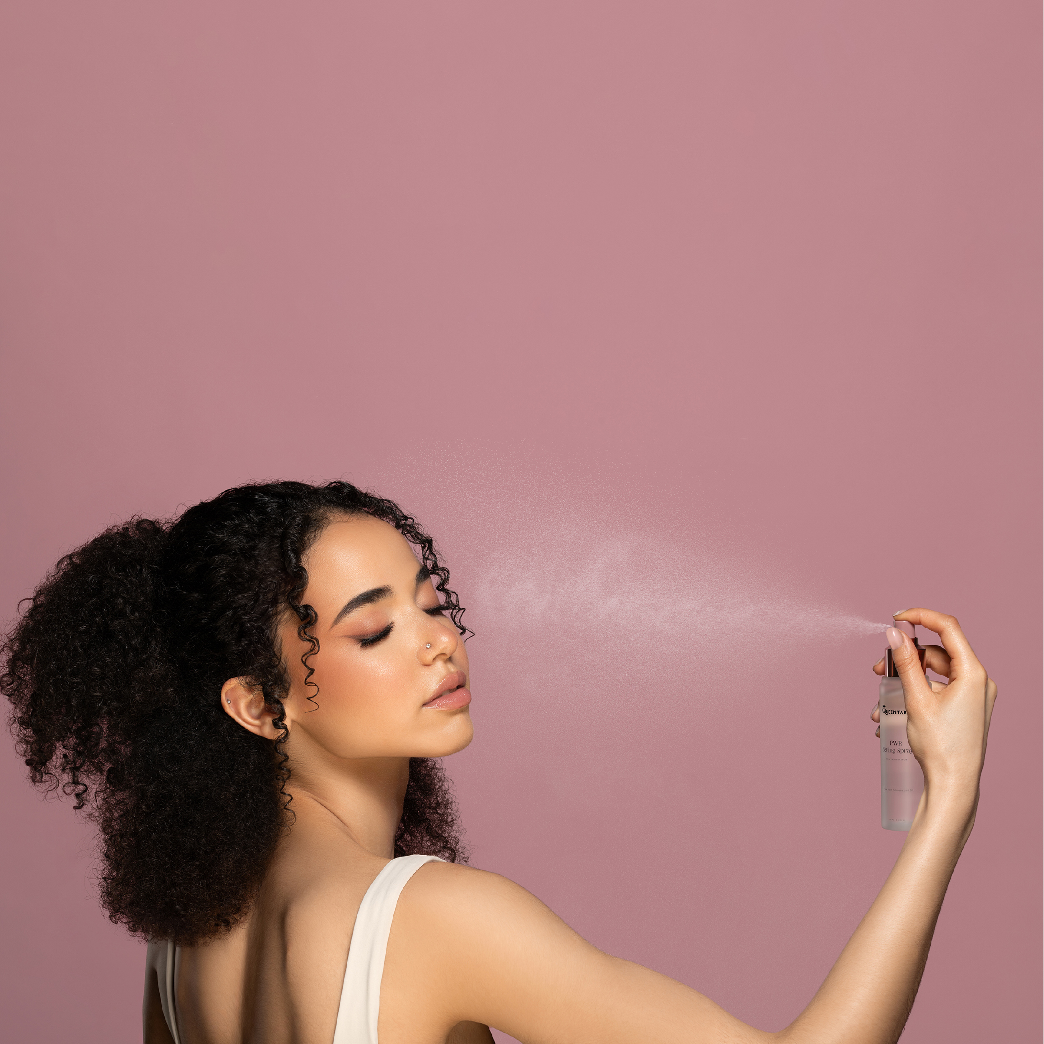 Vrouw met krullend haar die PWR Setting Spray gebruikt op haar gezicht, roze achtergrond