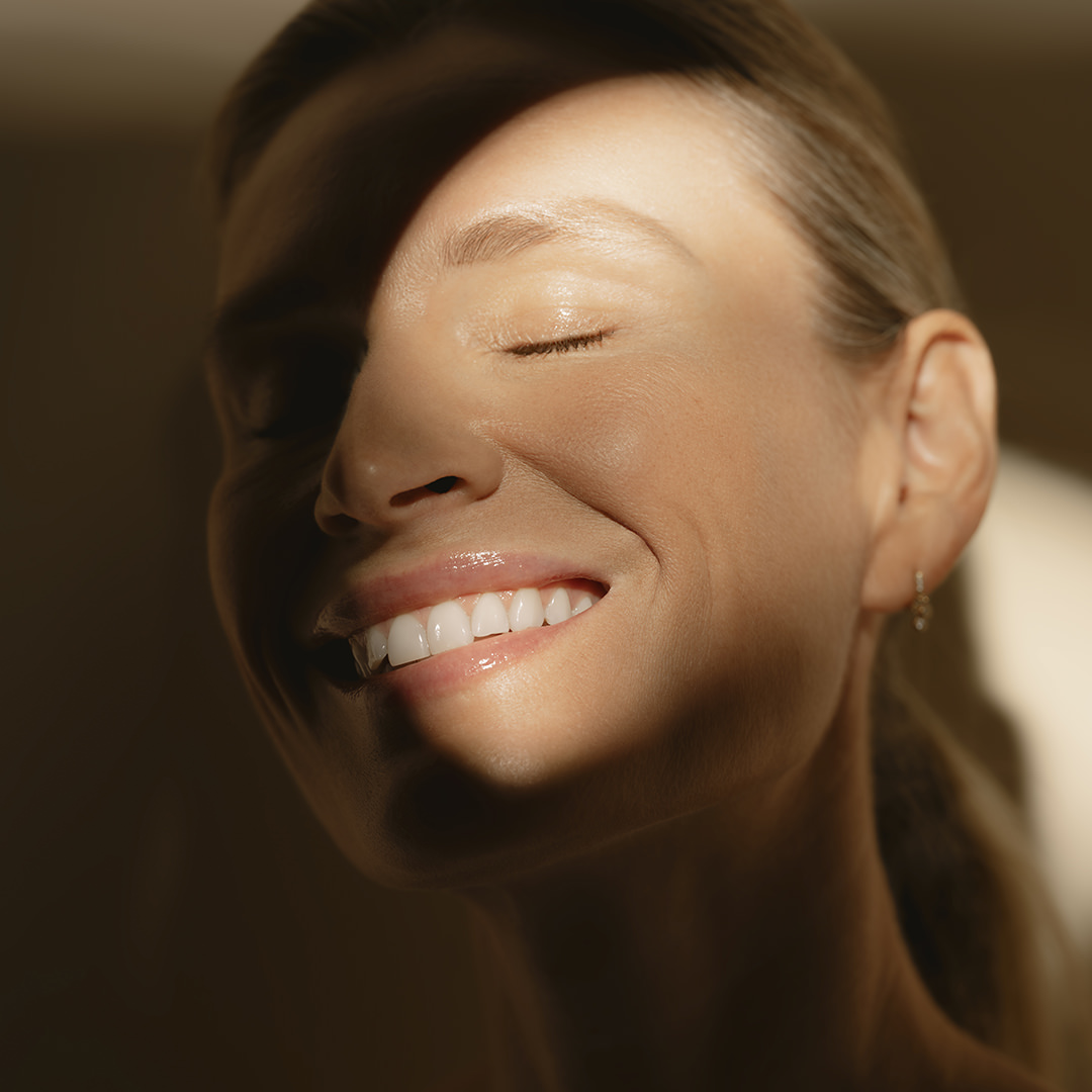 Close-up of a smiling woman with closed eyes, bathed in warm sunlight and shadows on her face