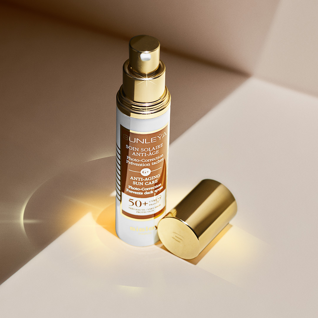 Sisley Sunleÿa SPF 50+ Anti-Aging Sun Care bottle with golden cap placed on a peach-toned surface under soft lighting