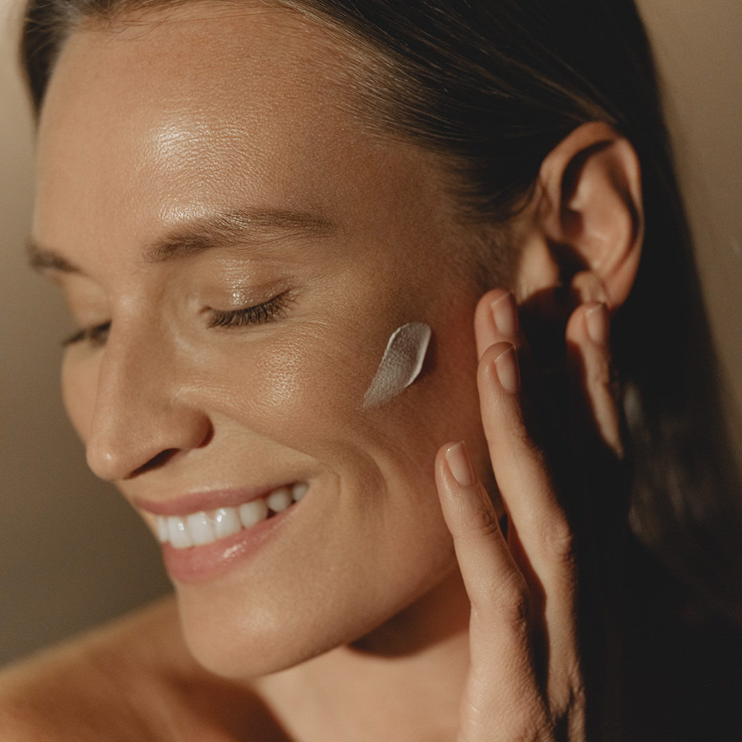Woman smiling gently while applying Sisley sunscreen to her cheek in warm, natural light